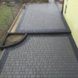 Ułożona szara kostka brukowa z ciemniejszym obramowaniem, widok z góry na podjazd, krawężnik z łukiem, w tle ogrodzenie i zaparkowany samochód.