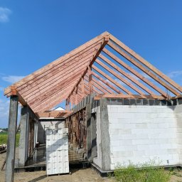 Ciesielstwo Artur Kościarz - Wznoszenie budynku z bloczków i drewnianej konstrukcji dachu na tle błękitnego nieba. Widoczne elementy konstrukcyjne i materiały budowlane.