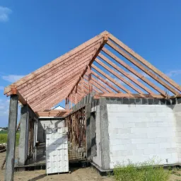 Wznoszenie budynku z bloczków i drewnianej konstrukcji dachu na tle błękitnego nieba. Widoczne elementy konstrukcyjne i materiały budowlane.