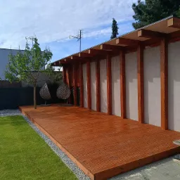 Nowoczesny taras drewniany z zadaszeniem i elementami betonowymi. Dwie wiszące huśtawki obok małego drzewa. Trawa i szara kostka brukowa.