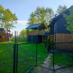 Nowoczesny dom z drewnianą altaną na podwyższeniu, widok zza zielonego ogrodzenia. W tle plac zabaw dla dzieci i drzewa.