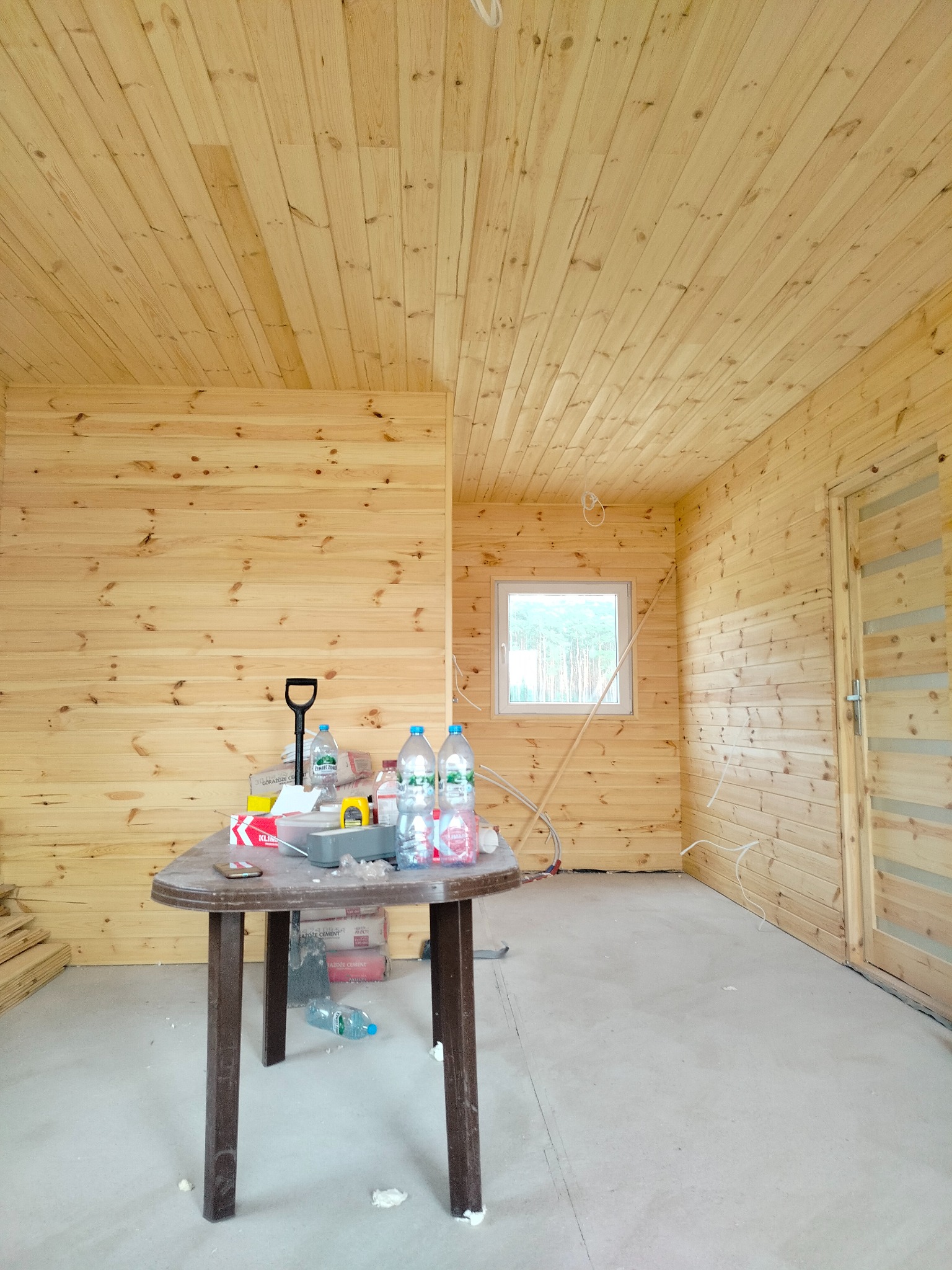 Wnętrze domu w trakcie wykończenia, ściany i sufit wyłożone drewnianą boazerią, widoczne okno z widokiem na las, stół z narzędziami i materiałami budowlanymi, betonowa podłoga.