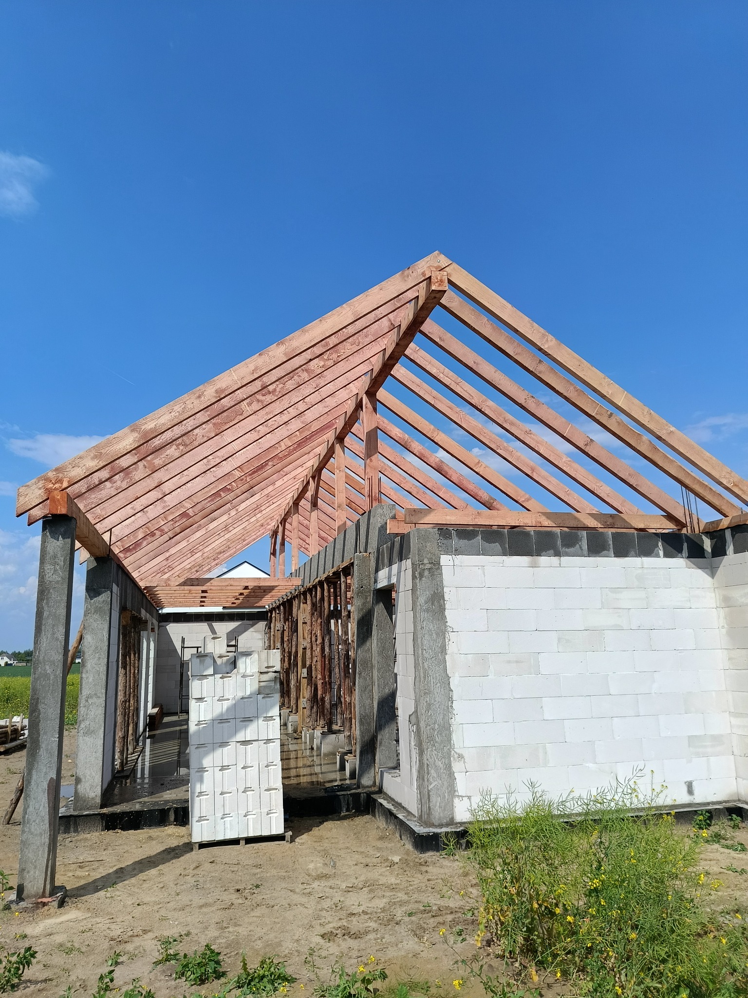 Wznoszenie budynku z bloczków i drewnianej konstrukcji dachu na tle błękitnego nieba. Widoczne elementy konstrukcyjne i materiały budowlane.