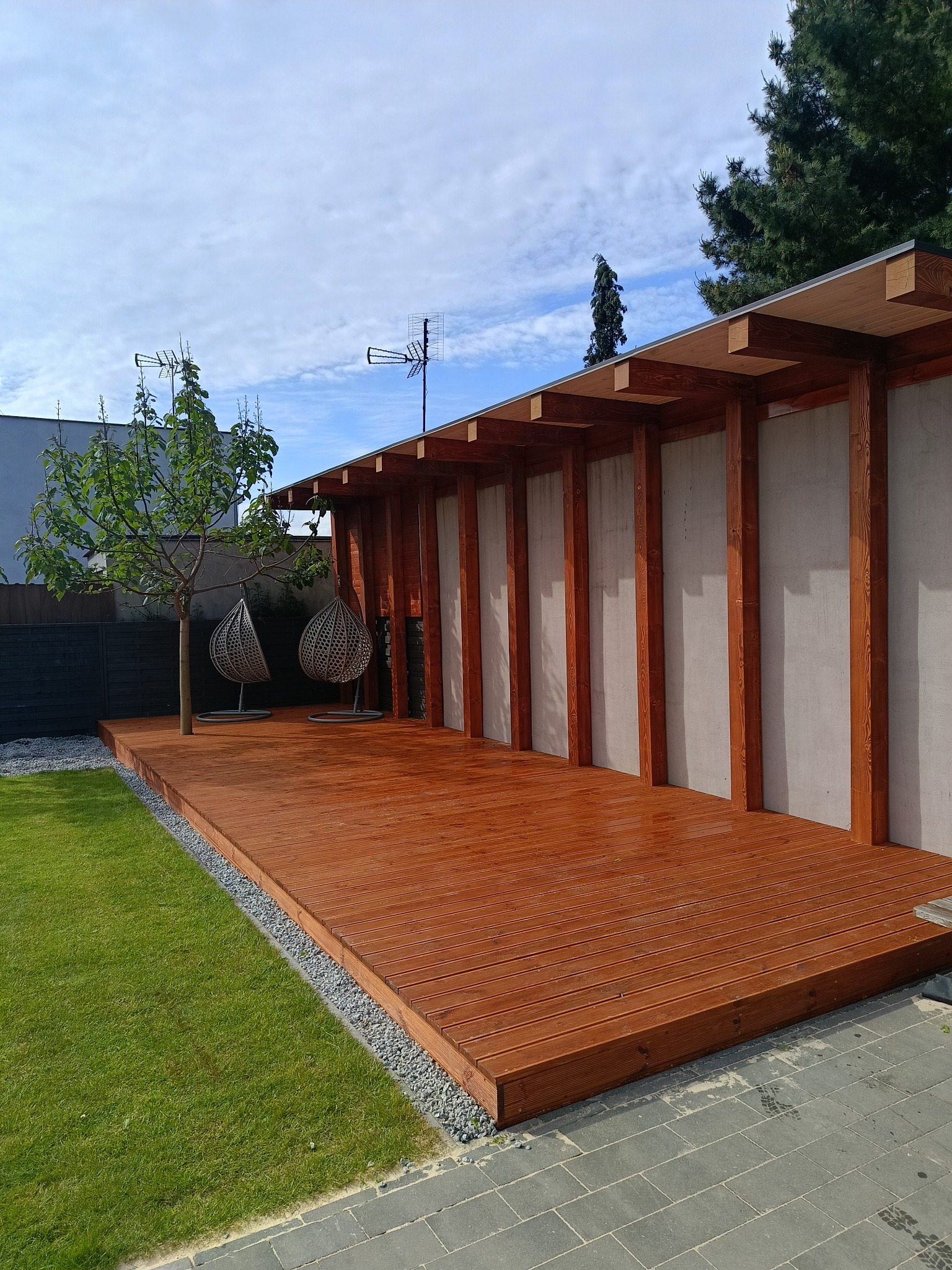 Nowoczesny taras drewniany z zadaszeniem i elementami betonowymi. Dwie wiszące huśtawki obok małego drzewa. Trawa i szara kostka brukowa.