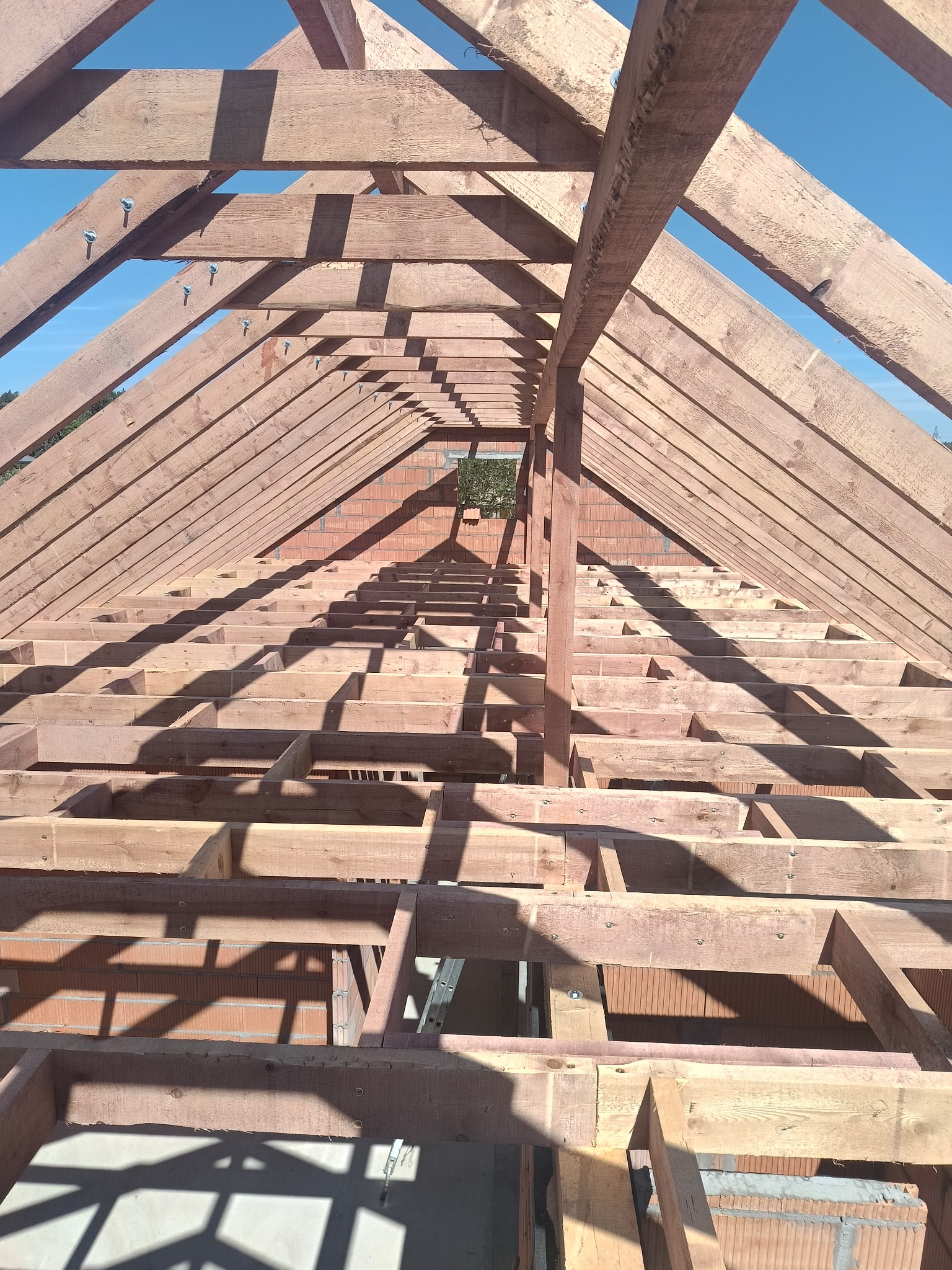 Drewniana konstrukcja dachu z widocznymi krokwiami i murłatami, połączonymi śrubami. W tle czerwona cegła i błękitne niebo. Ujęcie z dołu, w kierunku szczytu dachu.