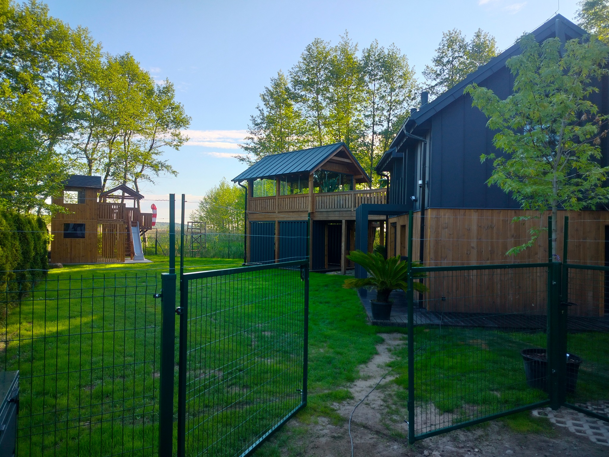 Nowoczesny dom z drewnianą altaną na podwyższeniu, widok zza zielonego ogrodzenia. W tle plac zabaw dla dzieci i drzewa.