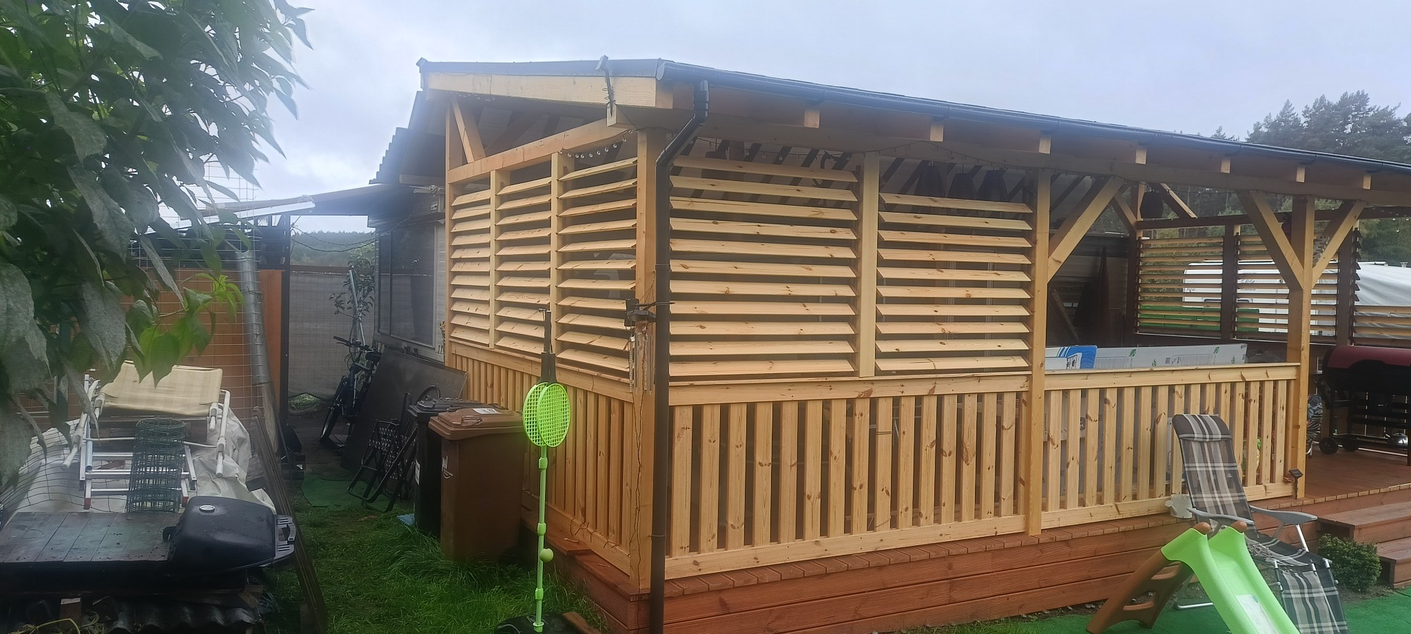 Drewniana altana z lamelami i barierką, widok z zewnątrz. Elementy konstrukcyjne z jasnego drewna, w tle zieleń. Na pierwszym planie zabawki i sprzęty ogrodowe.