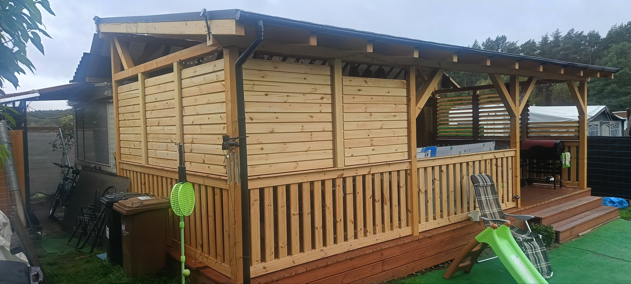 Drewniana altana z płaskim dachem i ażurowymi ścianami, otoczona zielenią. Widoczne detale konstrukcyjne i elementy wyposażenia.