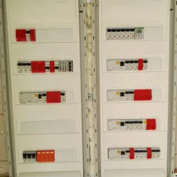 Dwa panele elektryczne z bezpiecznikami i przewodami zamontowane na surowej ścianie z betonu komórkowego, widoczne białe rurki instalacyjne na ścianie i pianka montażowa między panelami.