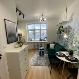 Aranżacja małego mieszkania: zielona sofa, okrągły stolik z dwoma nakryciami, komoda z obrazem leśnym i tapeta w liście.