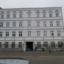 Odnowiona fasada kamienicy w trakcie prac wykończeniowych, widoczne rusztowania i pracownicy w jaskrawych kamizelkach, obok stara kamienica z anteną satelitarną.