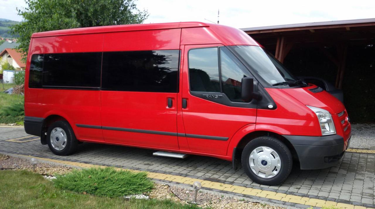 Czerwony bus Ford Transit z przyciemnianymi szybami, zaparkowany na kostce brukowej przed garażem, widoczny stopień wejściowy.