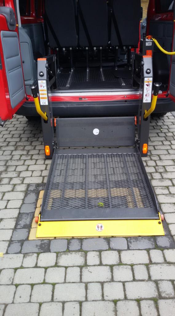 Czerwony bus z otwartymi drzwiami bagażnika i rozłożoną platformą dla osób niepełnosprawnych, widok z tyłu na brukowaną powierzchnię.