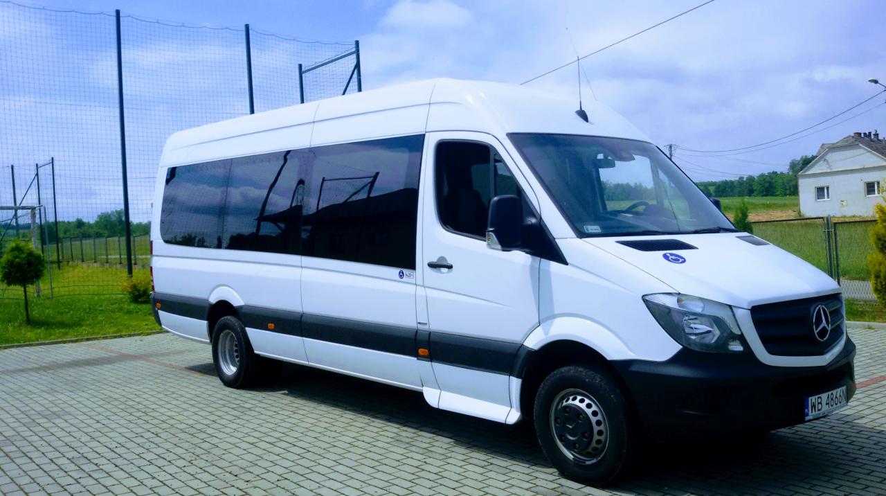 Biały bus Mercedes Sprinter z przyciemnianymi szybami, przystosowany do przewozu osób niepełnosprawnych, zaparkowany na kostce brukowej, w tle ogrodzenie boiska sportowego i budynek mieszkalny.