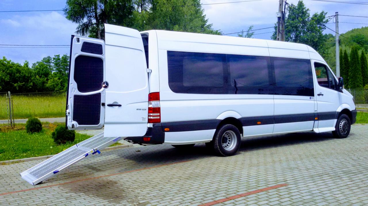 Biały bus Mercedes Sprinter z otwartymi tylnymi drzwiami i rozłożoną rampą dla wózków inwalidzkich, zaparkowany na brukowanym placu.