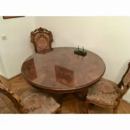 Okrągły, lakierowany stół z trzema krzesłami o tapicerowanych siedziskach i oparciach w stylu retro, na parkiecie, z zielonym przybornikiem na długopisy na blacie. Widoczne odbicia na powierzchni...