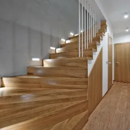 Drewniane schody z oświetleniem punktowym w nowoczesnym wnętrzu, betonowa ściana w tle, jasne poręcze i drewniane drzwi.