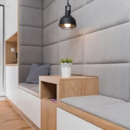 Wnęka z siedziskiem i tapicerowanym panelem ściennym, oświetlona wiszącą lampą, z drewnianym blatem i białą szafką, uzupełniona poduszką i doniczką z rośliną.
