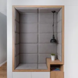 Wnęka w ścianie wykończona tapicerowanymi panelami w kolorze szarym, z drewnianą ramą i półką, oświetlona wiszącą lampą, z rośliną doniczkową i czasopismami.