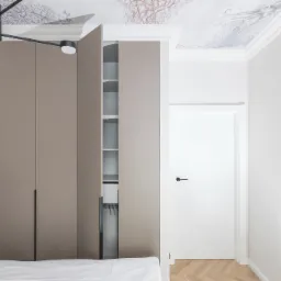 Beżowa szafa z otwartymi drzwiami w sypialni z parkietem i sufitem z motywem ośmiornicy, oświetlona nowoczesną lampą.