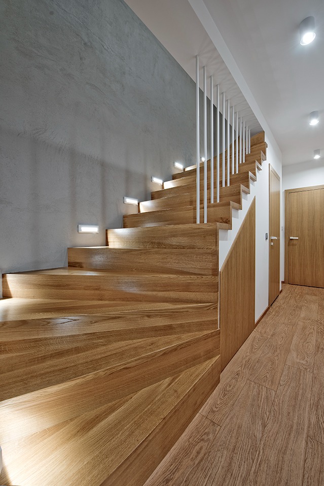 Drewniane schody z oświetleniem punktowym w nowoczesnym wnętrzu, betonowa ściana w tle, jasne poręcze i drewniane drzwi.