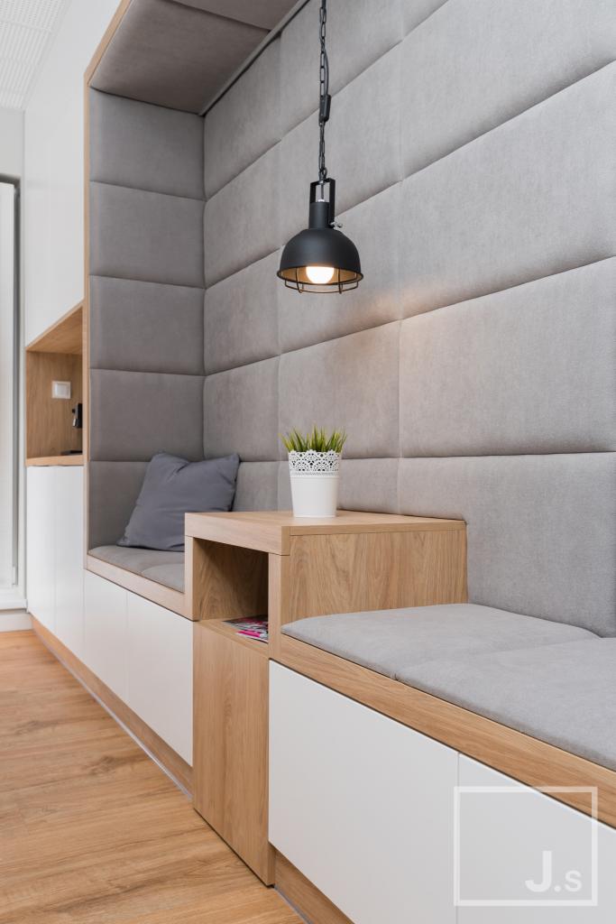 Wnęka z siedziskiem i tapicerowanym panelem ściennym, oświetlona wiszącą lampą, z drewnianym blatem i białą szafką, uzupełniona poduszką i doniczką z rośliną.