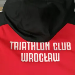 Czerwono-czarna bluza z kapturem, z białym haftem 'TRIATHLON CLUB WROCŁAW' na plecach. Widok z tyłu, detale haftu i kolorystyki.