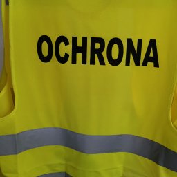 DERT Paweł Kwiatkowski - Żółta kamizelka odblaskowa z napisem 'OCHRONA' i szarymi pasami, zawieszona na wieszaku. Widoczne elementy wyposażenia pracowni.
