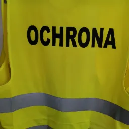 Żółta kamizelka odblaskowa z napisem 'OCHRONA' i szarymi pasami, zawieszona na wieszaku. Widoczne elementy wyposażenia pracowni.