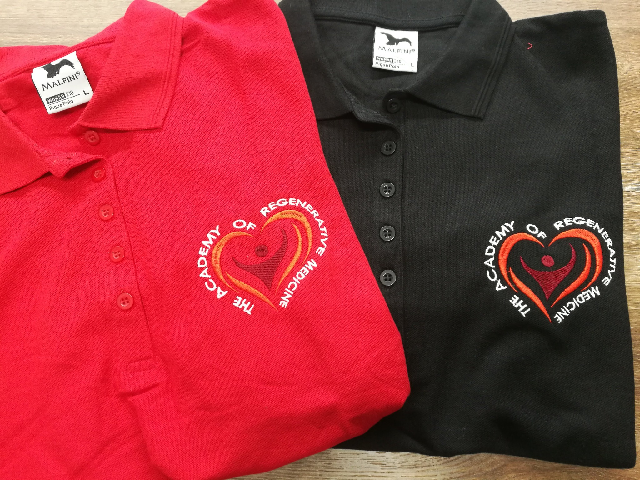 Czerwona i czarna koszulka polo z haftowanym logo 'The Academy of Regenerative Medicine', metki Malfini, rozmiar L, leżące na drewnianej powierzchni.
