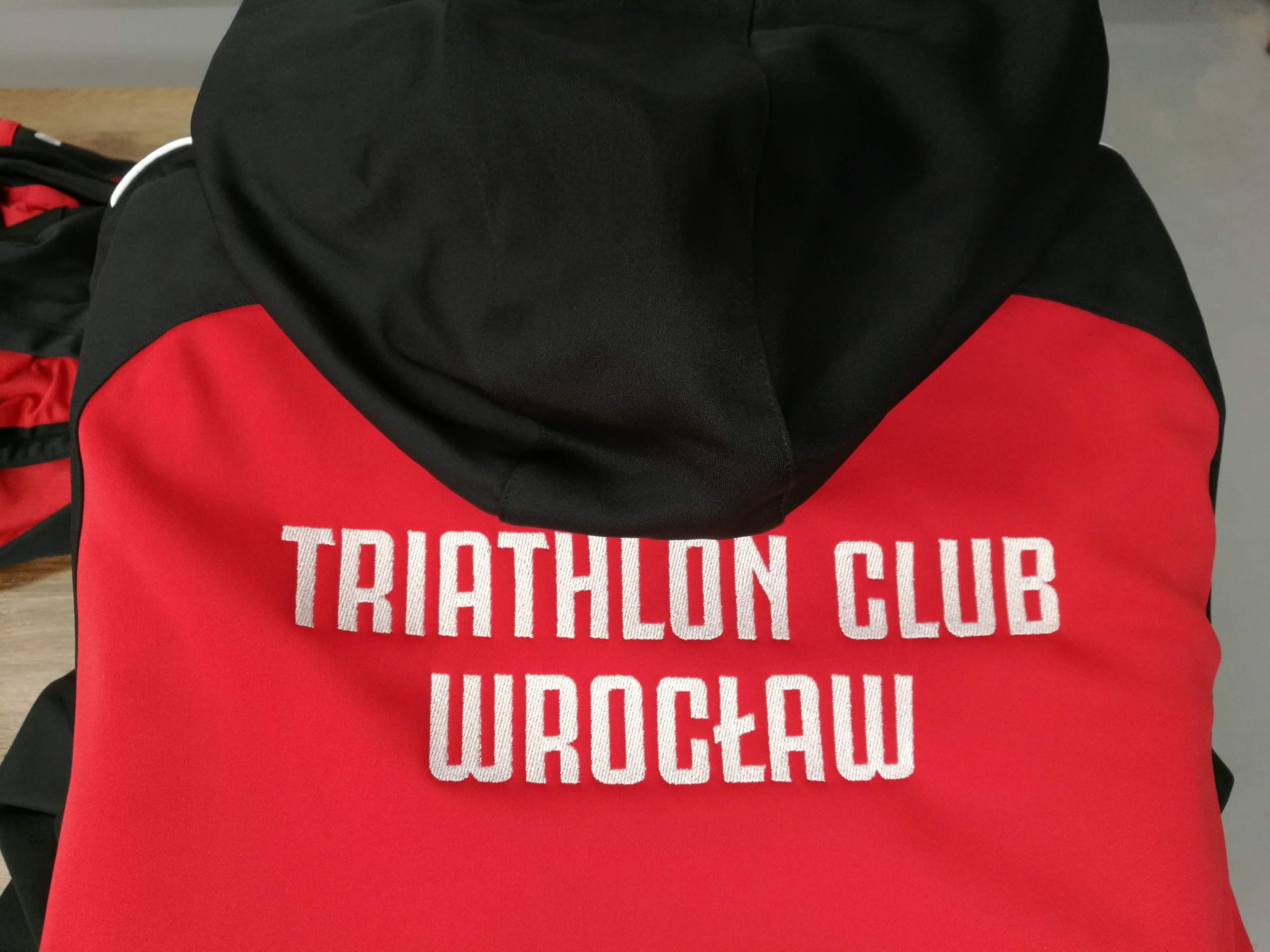 Czerwono-czarna bluza z kapturem, z białym haftem 'TRIATHLON CLUB WROCŁAW' na plecach. Widok z tyłu, detale haftu i kolorystyki.