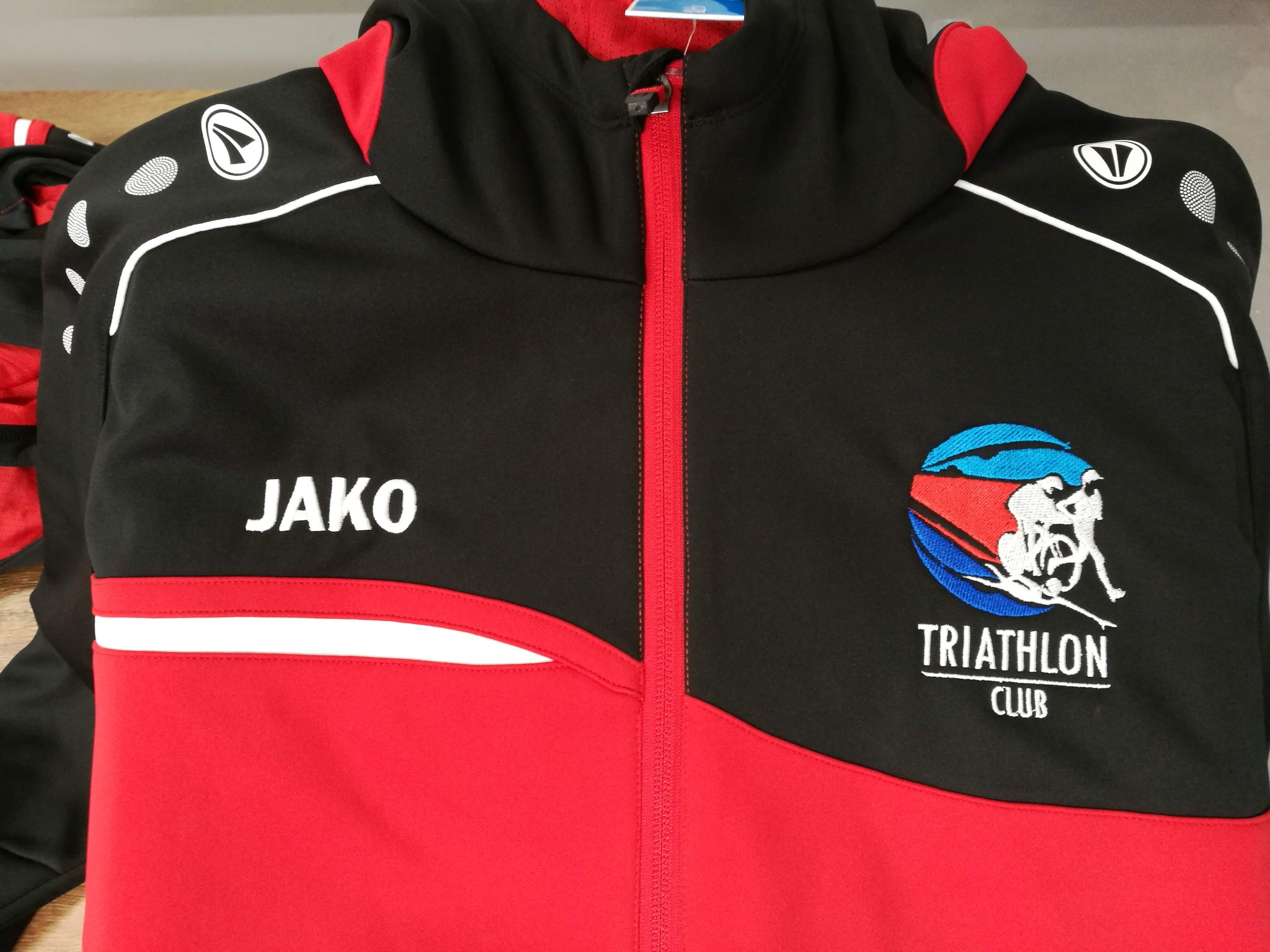 Czarno-czerwona bluza sportowa JAKO z haftowanym logo klubu triathlonowego. Widoczny zamek błyskawiczny i detale wykończenia.