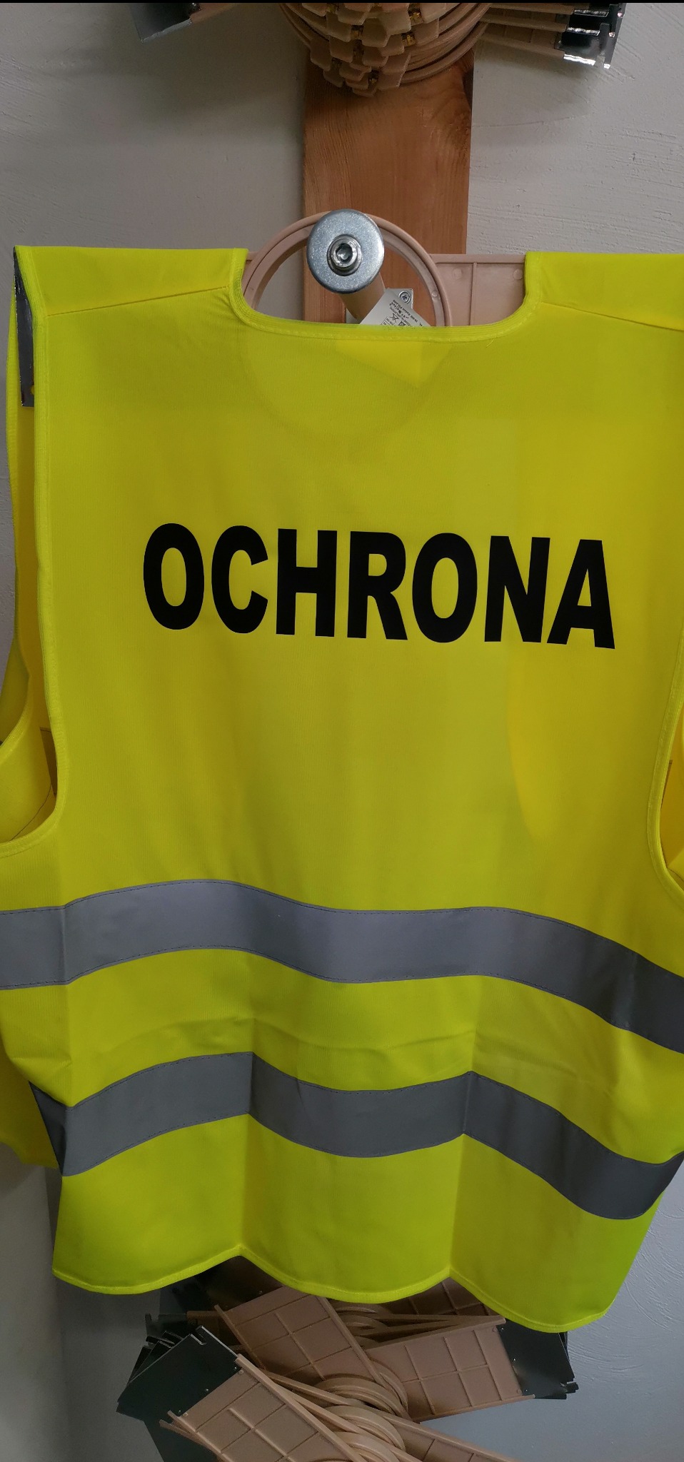 Żółta kamizelka odblaskowa z napisem 'OCHRONA' i szarymi pasami, zawieszona na wieszaku. Widoczne elementy wyposażenia pracowni.
