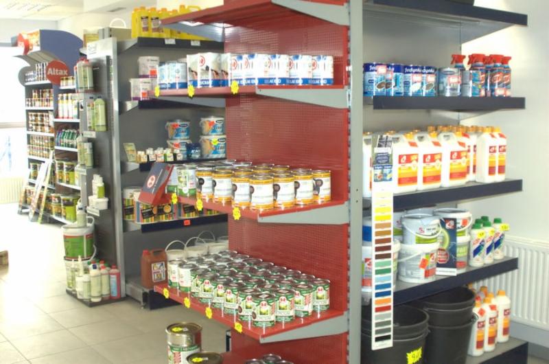 Wnętrze sklepu w Knurowie z półkami wypełnionymi farbami, lakierami i innymi produktami do wykończenia wnętrz. Widoczne puszki, wiadra i butelki różnej wielkości, a także paleta kolorów.