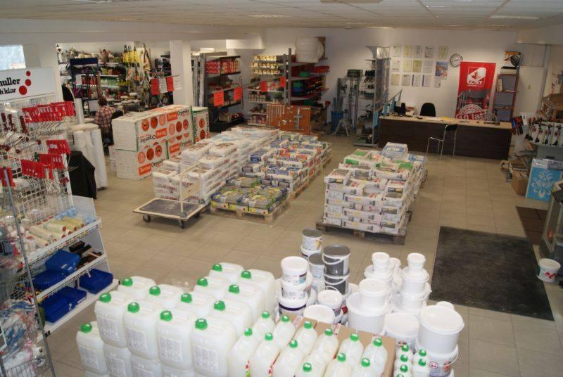 Wnętrze sklepu z materiałami budowlanymi, widok z góry na alejki z paletami wypełnionymi workami i kartonami, w tle regały z narzędziami i farbami, a w głębi biurko obsługi klienta.