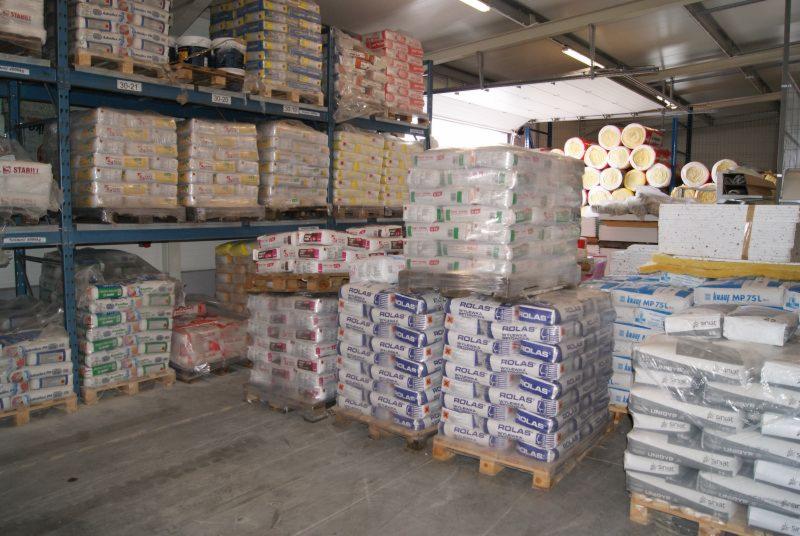 Wnętrze magazynu z materiałami budowlanymi ułożonymi na paletach i regałach, widoczne worki z zaprawami, klejami i materiałami izolacyjnymi, oświetlenie sufitowe.