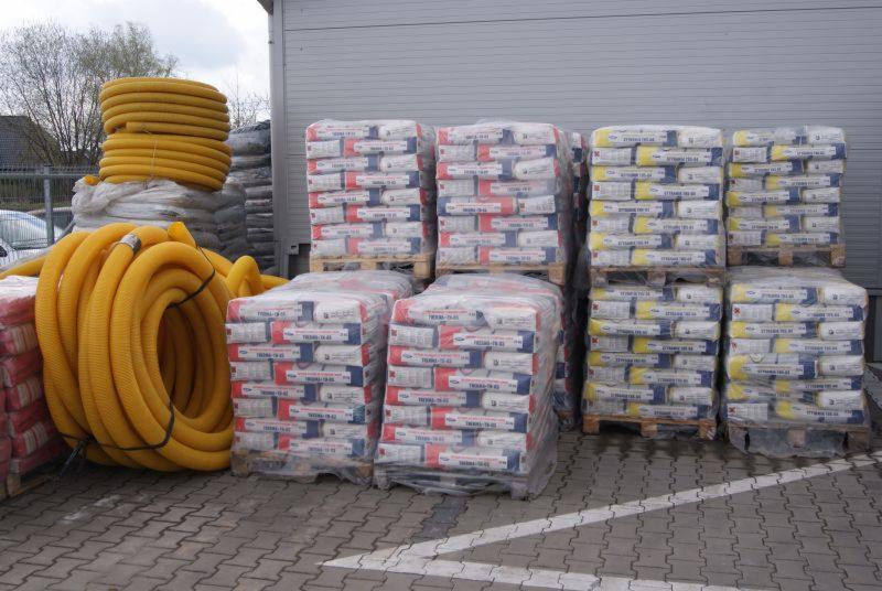 Stosy worków z zaprawą murarską na paletach, obok zwinięte żółte rury drenażowe, na tle szarej hali magazynowej.