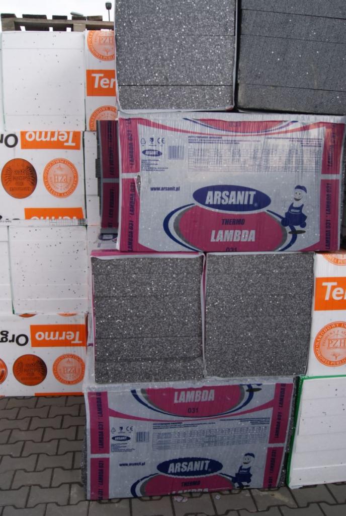 Ułożone na palecie paczki szarego i białego materiału termoizolacyjnego, owinięte folią z nadrukiem nazwy producenta i parametrami technicznymi.