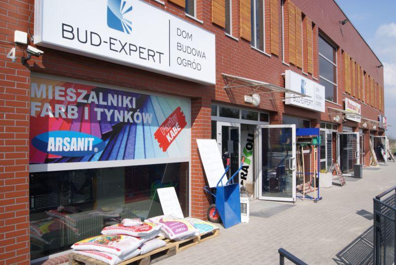 Fasada sklepu budowlanego BUD-EXPERT z oknem wystawowym prezentującym mieszalniki farb i tynków Arsanit, worki z materiałami na palecie przed wejściem oraz niebieski wózek budowlany.