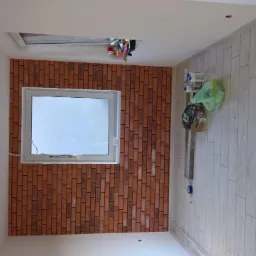 Wnętrze pokoju w trakcie wykańczania: ściana z cegiełką przy oknie, białe panele podłogowe, widoczne akcesoria i narzędzia budowlane.