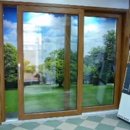 Dwuskrzydłowe drzwi przesuwne z drewnopodobną ramą, prezentowane w salonie wystawowym na tle plakatu z widokiem zielonego ogrodu i błękitnego nieba; obok ekspozytor z próbkami wykończeń.