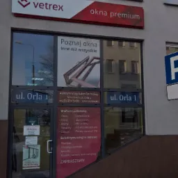 Fasada budynku z witryną salonu sprzedaży stolarki okiennej i drzwiowej Vetrex, widoczne plakaty reklamowe oraz tablica informacyjna z ofertą firmy, znak zakazu parkowania na pierwszym planie.