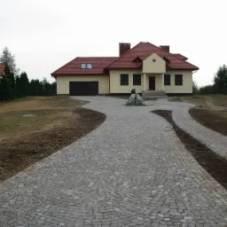 Posesja z podjazdem z kostki granitowej ułożonej we wzór wachlarza. Na podjeździe kamienna dekoracja. W tle dom z brązowymi oknami i czerwoną dachówką.