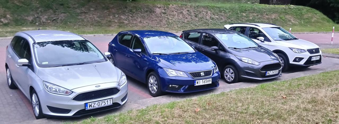 Rząd czterech samochodów różnych marek i kolorów zaparkowanych obok siebie na parkingu, widocznych od przodu.