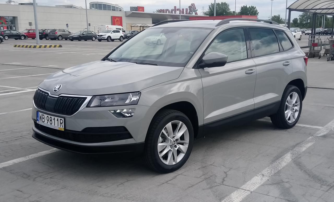 Szary SUV Skoda Karoq zaparkowany na parkingu przed centrum handlowym w pochmurny dzień.