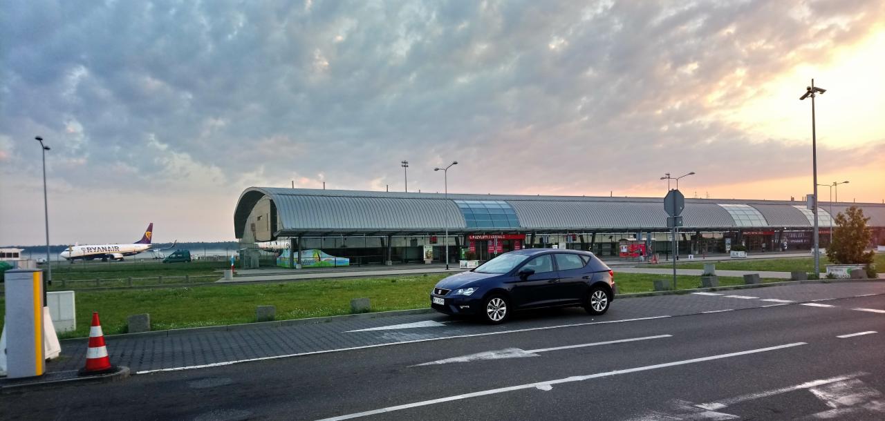 Granatowy samochód osobowy przed terminalem lotniska w Warszawie, na drugim planie samolot Ryanair i zachmurzone niebo o zmierzchu.