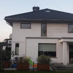 Dwupiętrowy dom jednorodzinny z czarnym dachem, kremową elewacją i drewnianymi oknami, widoczny z ogrodu z trawnikiem i roślinami w drewnianych donicach.