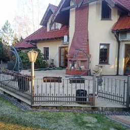 Przestronny taras z kamienną nawierzchnią, otoczony ozdobnym, metalowym ogrodzeniem, z zewnętrznym kominkiem z czerwonej cegły i wiszącym fotelem bujanym, w tle dom z czerwoną dachówką.