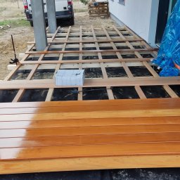 SHINE WOOD - Budowa tarasu: widok na drewniany szkielet konstrukcji, częściowo pokryty deskami z widocznymi narzędziami i opakowaniem, na podłożu z czarnej membrany, z dostawczym busem w tle.