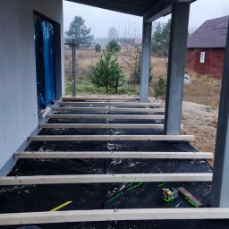 SHINE WOOD - Budowa tarasu: widok na legary ułożone na czarnej membranie, z widocznymi narzędziami i poziomnicą laserową, w tle dom w trakcie budowy i zalesiony krajobraz.